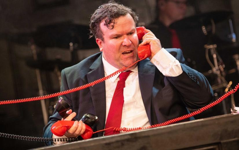 Actor playing Gordon Brown picking up red telephones in a black suit with red tie
